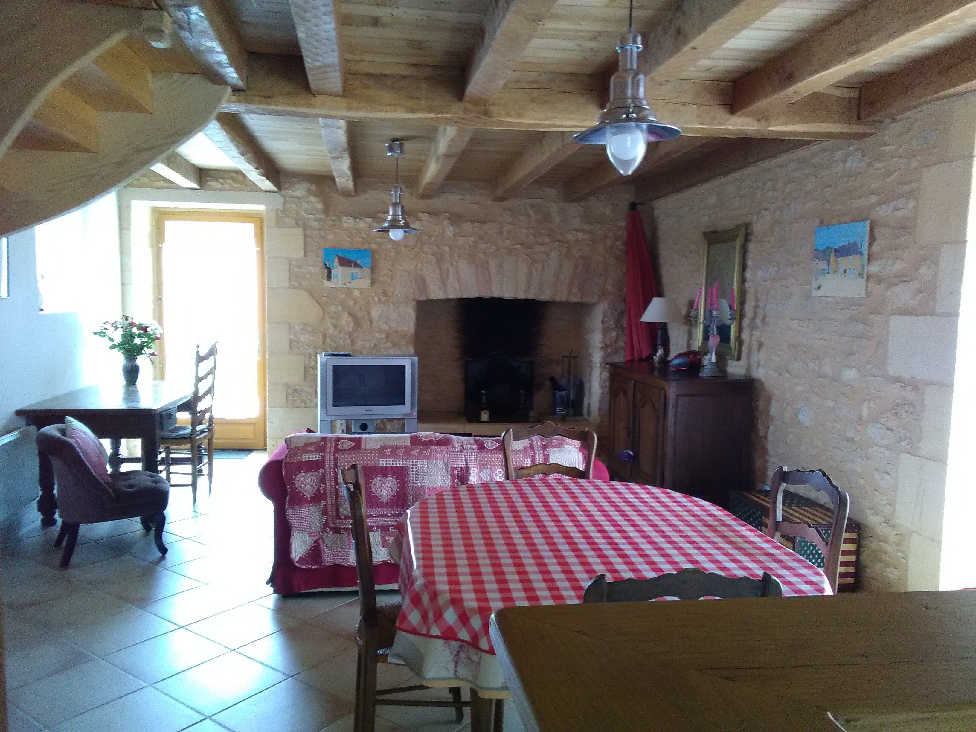 Une pièce rustique avec un mur en pierre, des poutres en bois, une cheminée, un téléviseur et un coin repas avec une nappe à carreaux.
