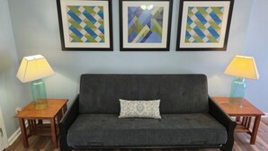 Una habitación con un sofá gris, dos mesitas de madera con lámparas, y tres cuadros geométricos enmarcados en la pared.
