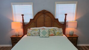 Un dormitorio con una cama grande, dos mesitas de noche con lámparas, un banco al pie de la cama y dos ventanas con persianas.
