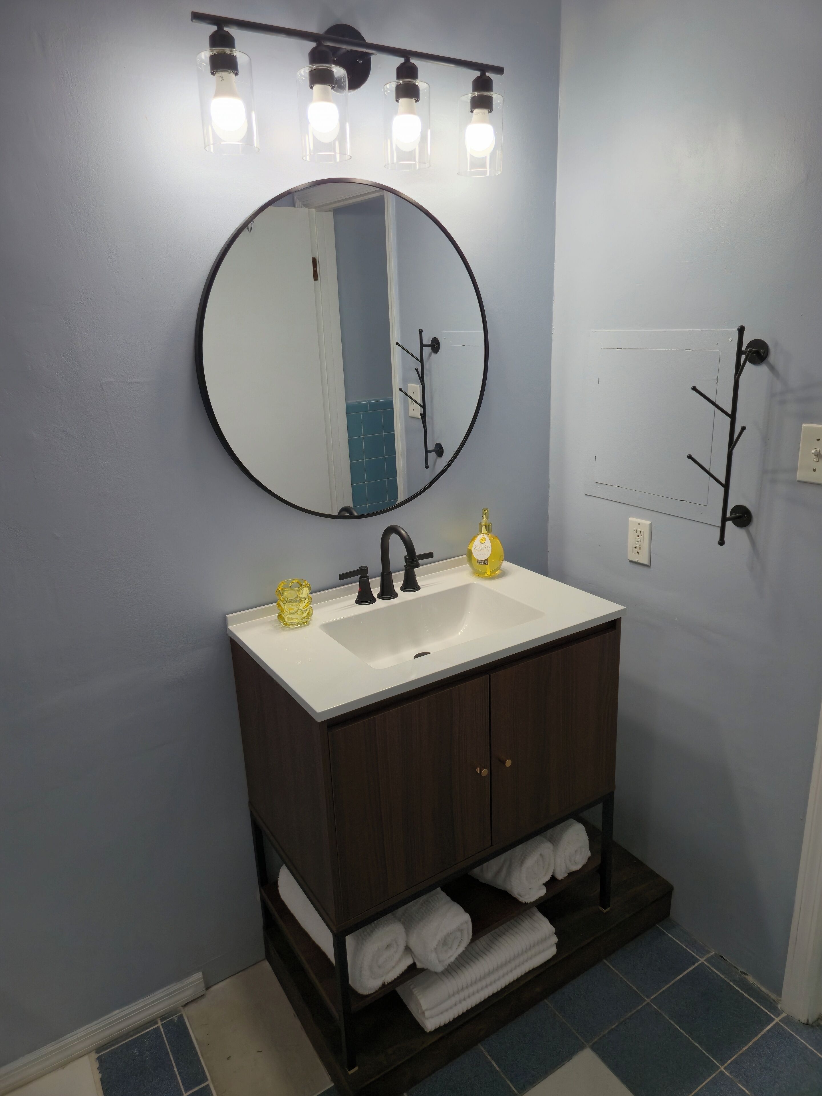 A bathroom with a round mirror, a white sink, and a dark wooden vanity. There are three light fixtures above the mirror and a towel rack on the wall.