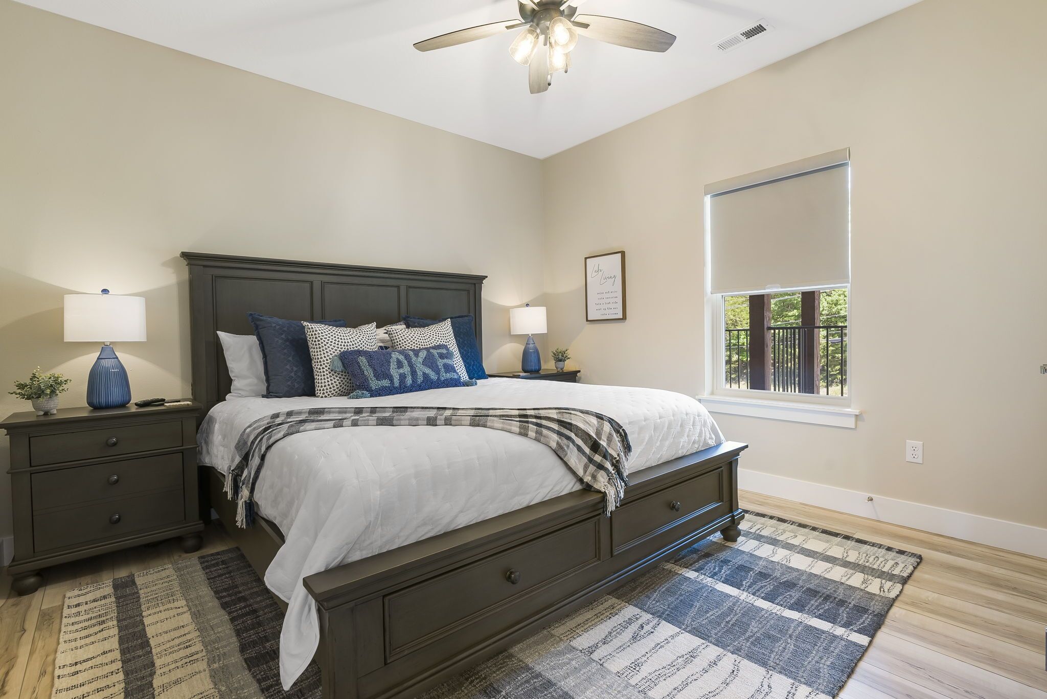 A bedroom with a dark wooden bed, two bedside tables with lamps, a large window with a view, and a ceiling fan.