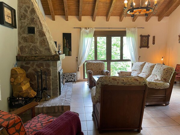 A cozy living room with a stone fireplace, wooden furniture, and large windows. - Casa Trasgu Para 8 Personas en Tornín, Cangas de Onís (Asturias)