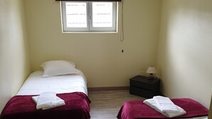 Ein Zimmer mit zwei Betten, jeweils mit weißer Bettdecke und roter Decke, einem kleinen Nachttisch mit Lampe und einem Fenster mit Jalousien.