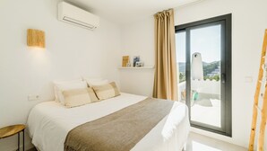 A bedroom with a bed, a bedside table, a wall-mounted air conditioner, a window with curtains, and a view of a building outside. - Casa Zita Mar - Ibiza style villa, beach nearby & magical view (Cala Llonga)