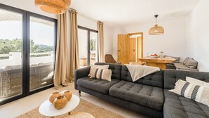 A modern living room with a grey sofa, a round dining table, and a large window offering a view of the outdoors. - Casa Zita Mar - Ibiza style villa, beach nearby & magical view (Cala Llonga)