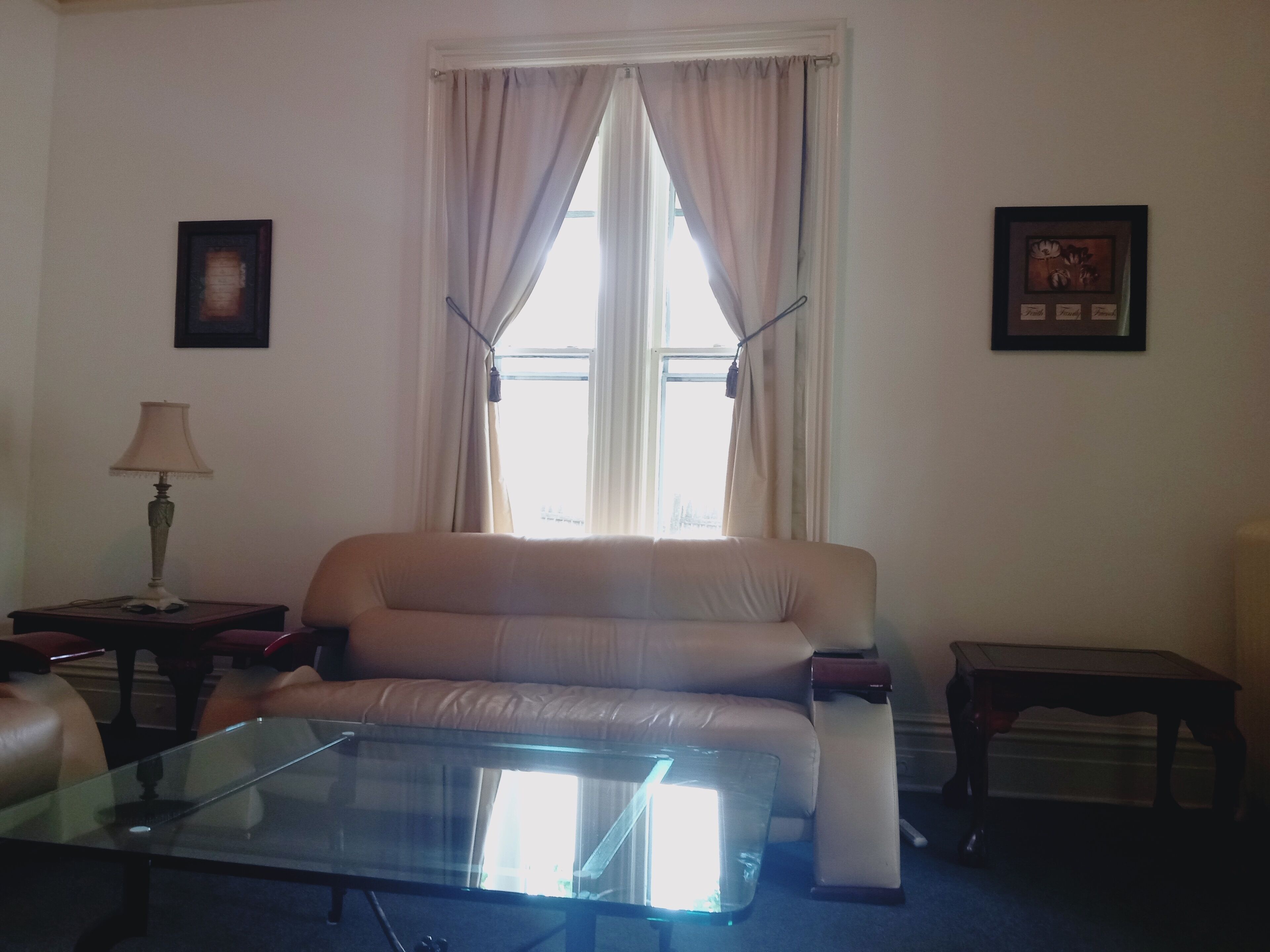 Ein Wohnzimmer mit einem beigefarbenen Sofa, einem Glastisch, einer Lampe und zwei gerahmten Bildern an den Wänden.