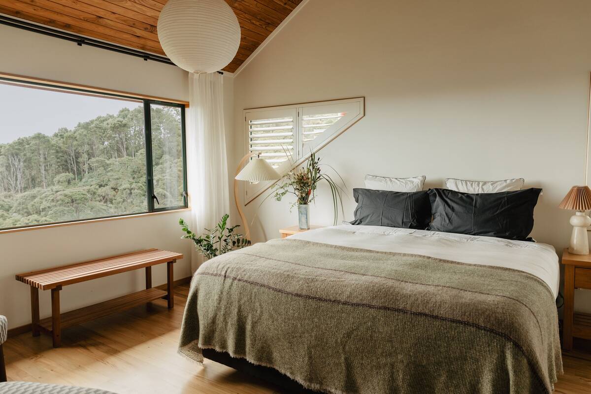 A bedroom with a large window, a wooden bench, a bed with a green blanket, and a bedside table with a lamp.