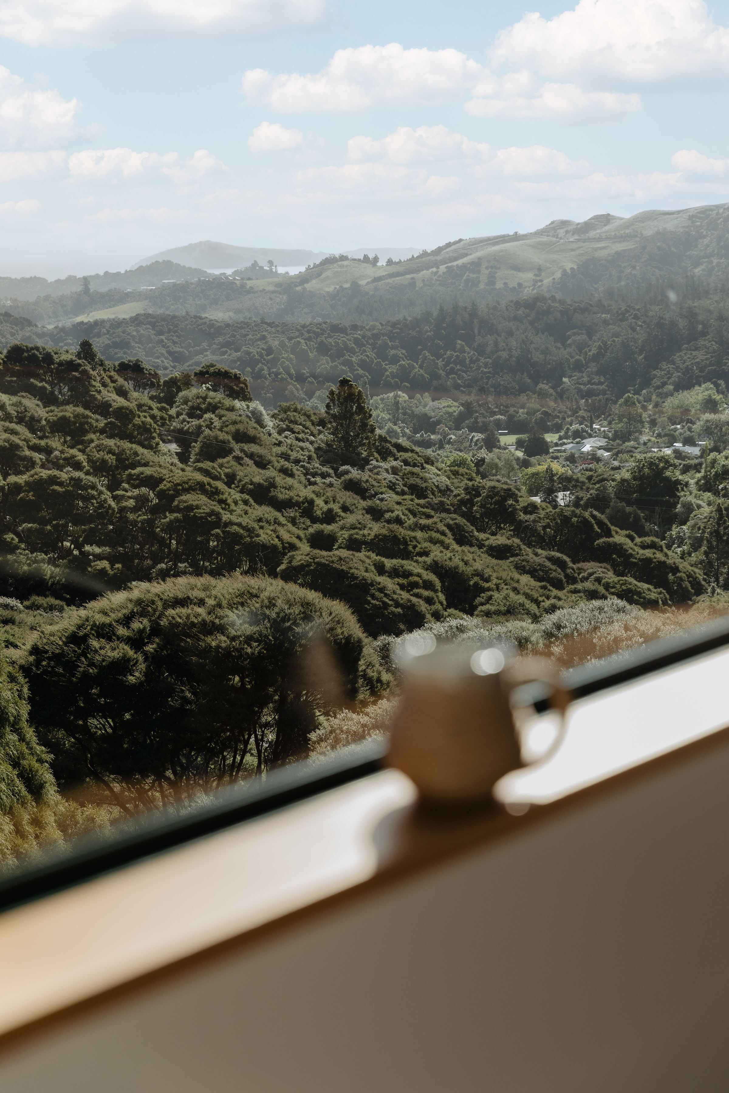 A view from a window overlooking a hilly landscape with dense greenery and a distant village.