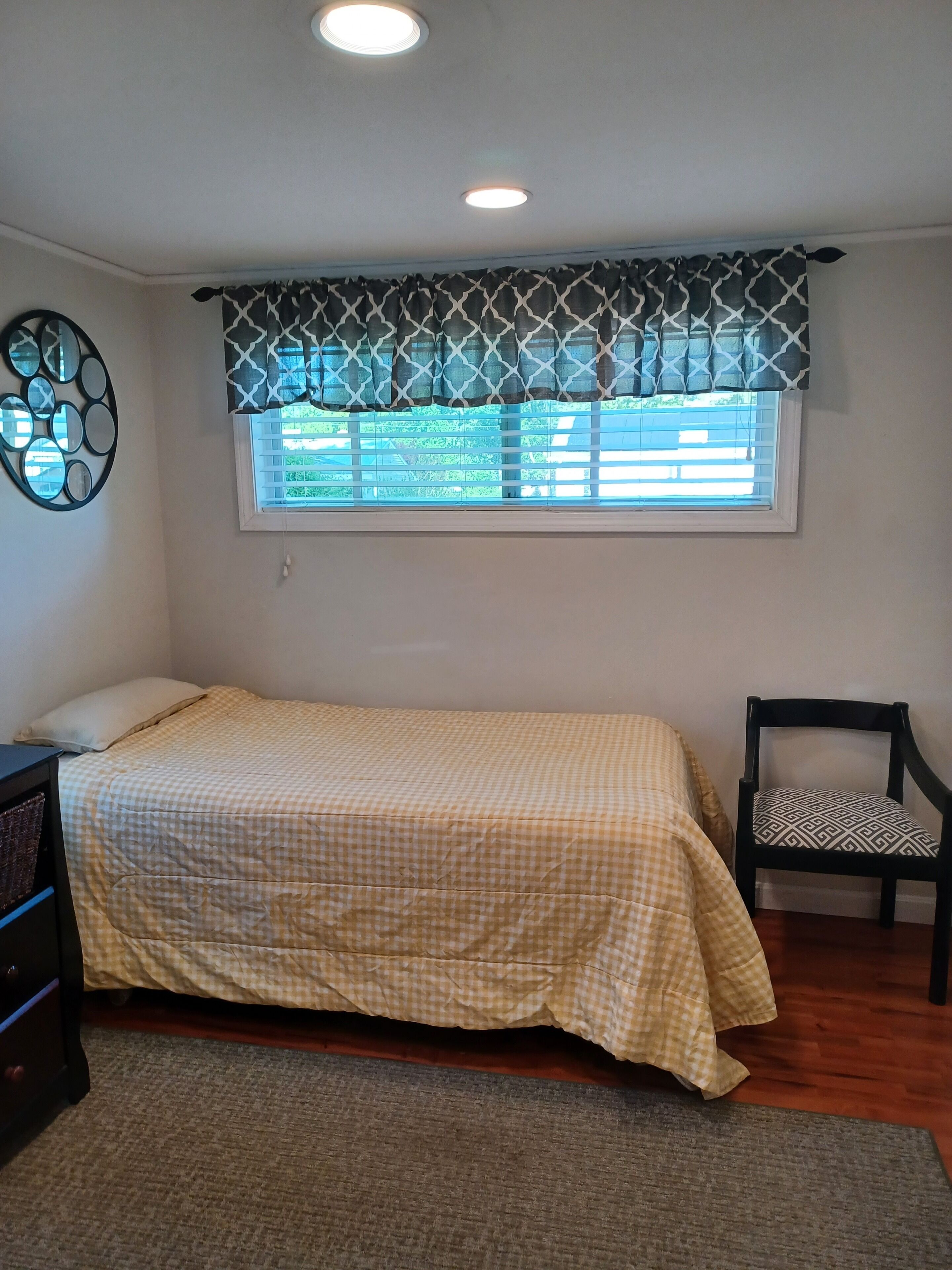 A bedroom with a bed, a chair, a dresser, and a window with patterned curtains.