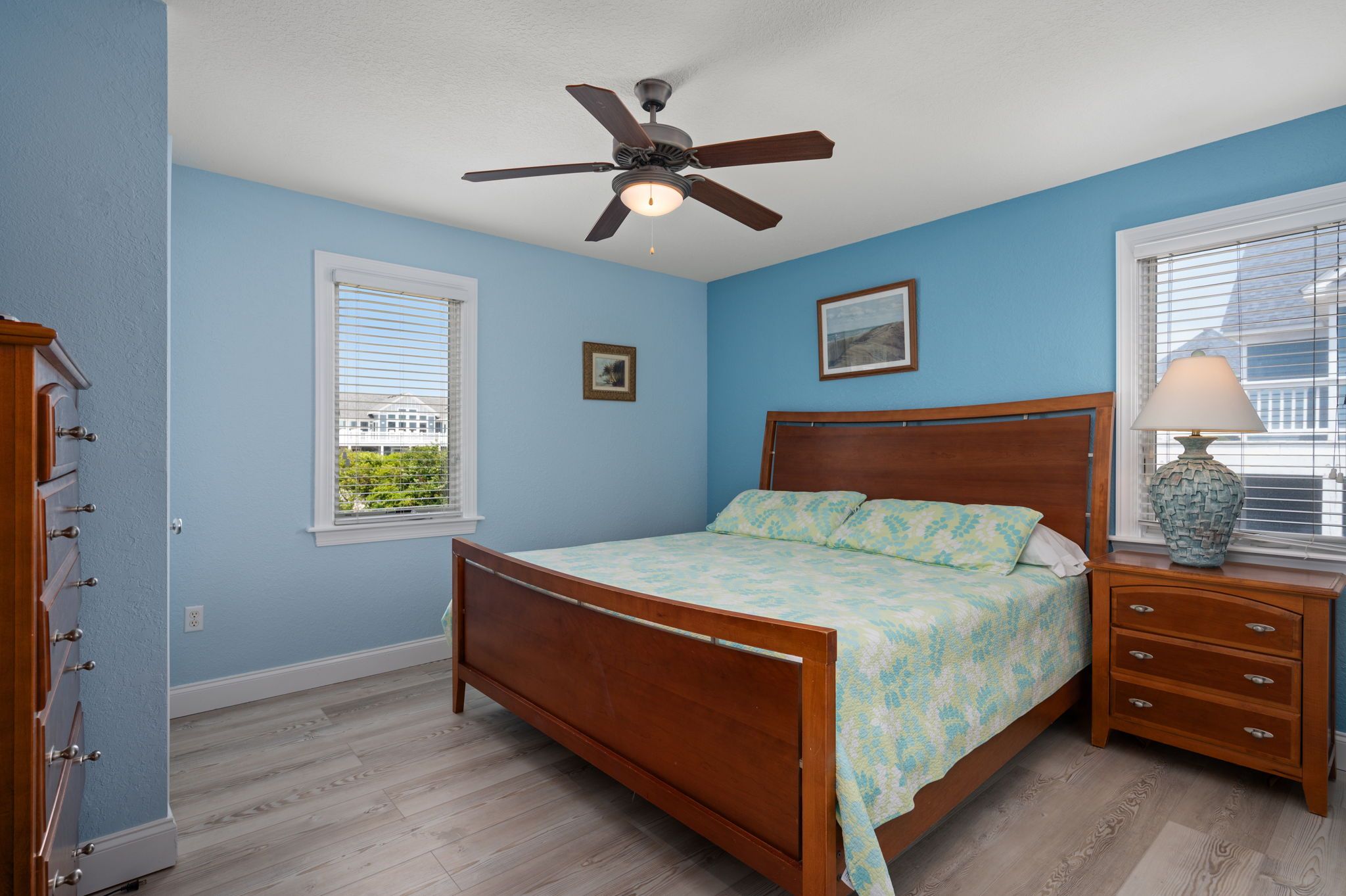 A bedroom with a wooden bed, a ceiling fan, a nightstand with a lamp, and a window with blinds.