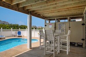 Zona de piscina techada con techo de madera, piscina, sillas de descanso y un cartel con las normas de la piscina.