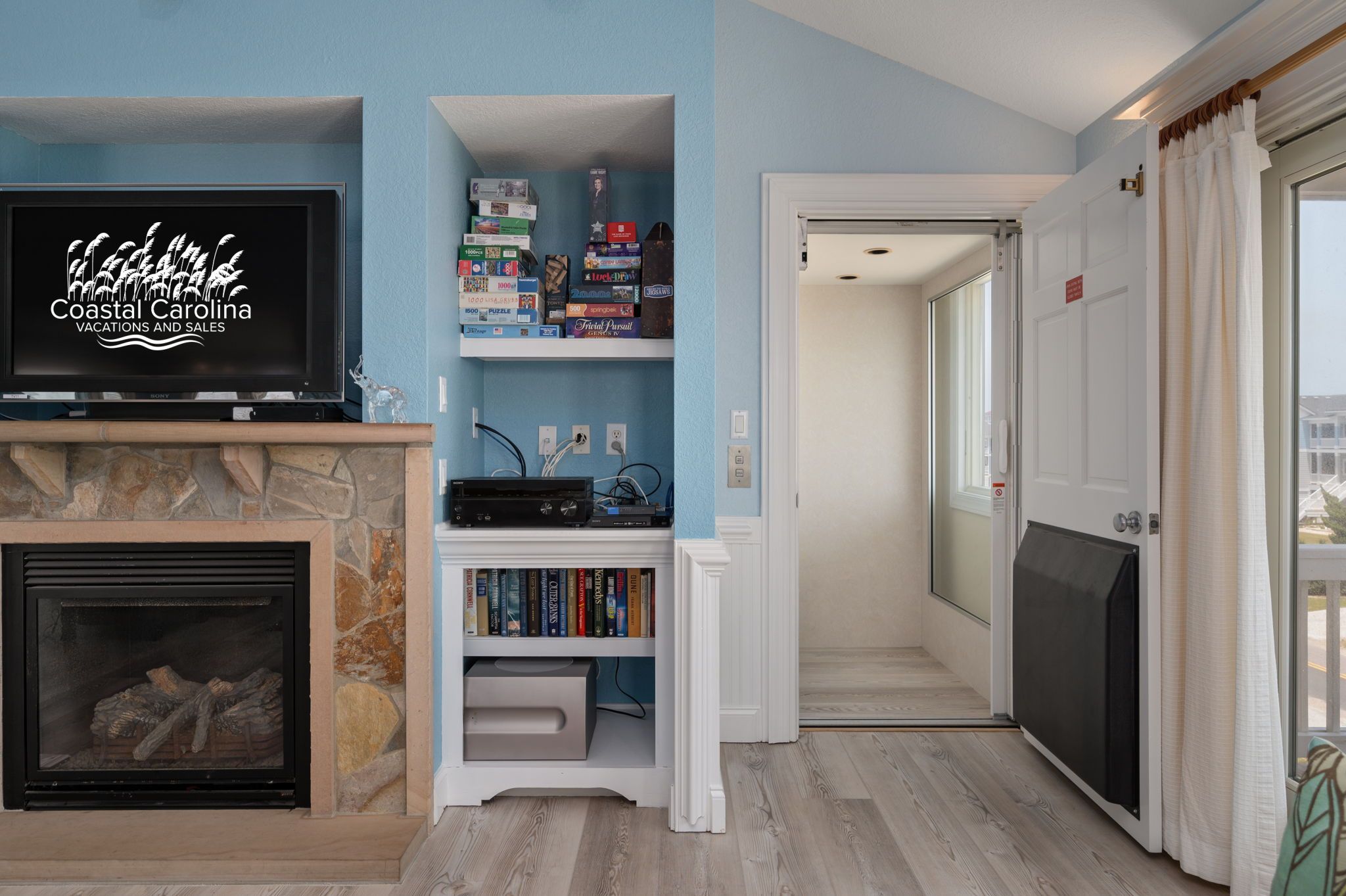 A living room with a stone fireplace, a television displaying 'Coastal Carolina Vacations and Sales', and a bookshelf with various items.