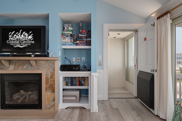 A living room with a stone fireplace, a television displaying 'Coastal Carolina Vacations and Sales', and a bookshelf with various items.
