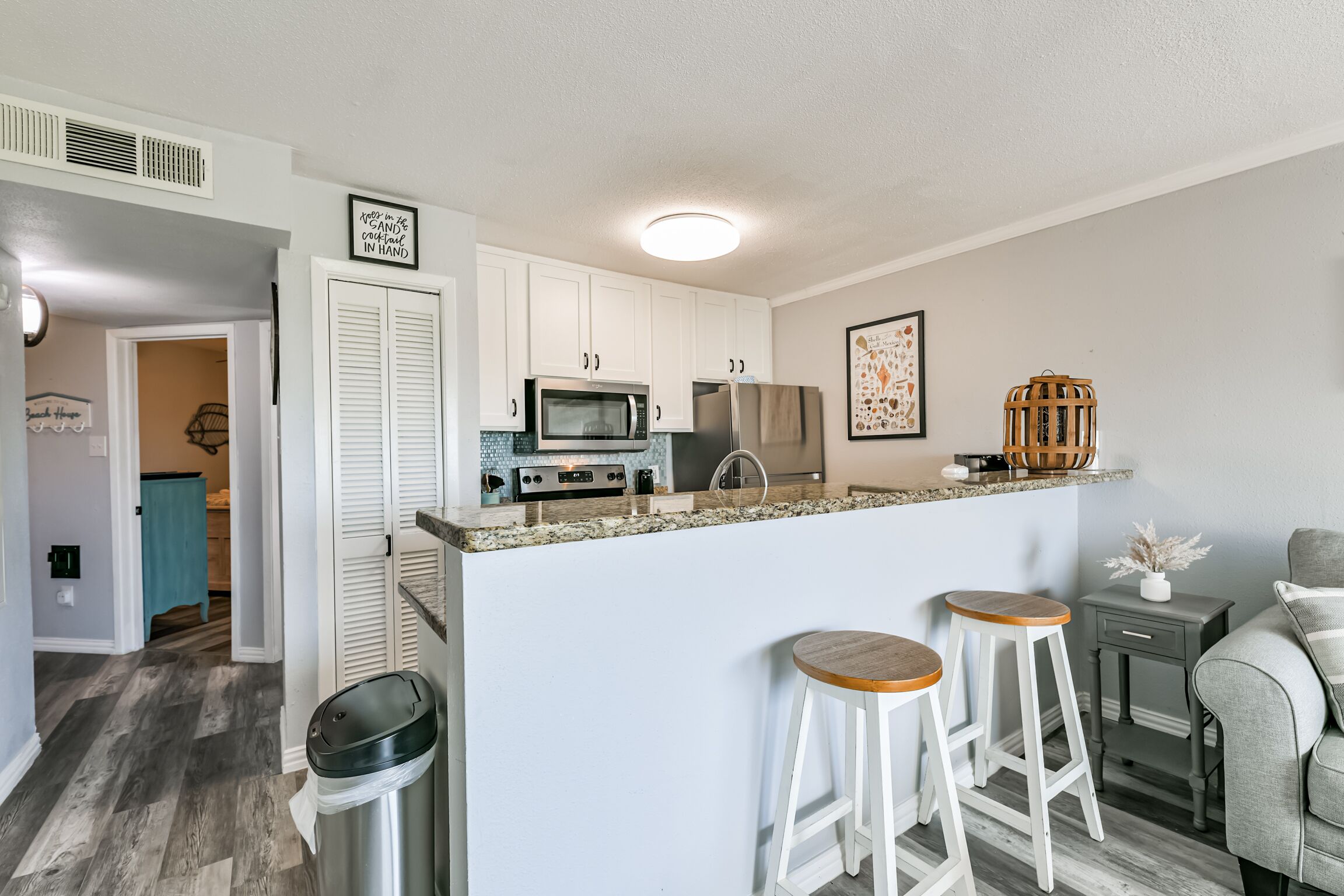 A compact kitchen with white cabinets, a granite countertop, and a microwave. There are two wooden stools and a small side table.
