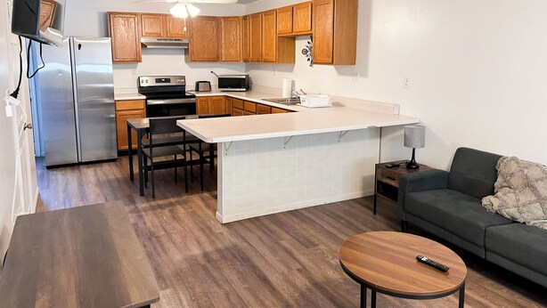 A compact kitchen with wooden cabinets, a refrigerator, a microwave, and a stove. There is a dining area with a table and chairs, and a small seating area with a sofa and a coffee table. - Cozy 2BR, 1BA Getaway in Macomb, IL (Macomb)