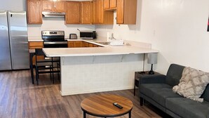 A modern kitchen with wooden cabinets, a stainless steel refrigerator, and a built-in island with a sink. There is a dining area with a round wooden table and two chairs. A gray sofa is in the living area with a remote control on it. - Cozy 2BR, 1BA Getaway in Macomb, IL (Macomb)