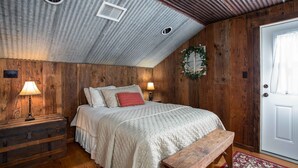 A bedroom with a wooden ceiling, a bed with a quilted blanket, a bench, a door, and a wall-mounted light fixture.