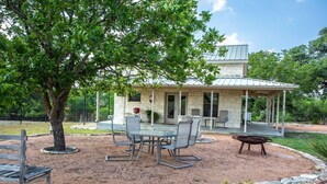 A covered patio with a stone building in the background, outdoor seating, and a tree in the foreground.