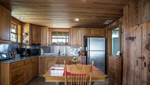 A rustic kitchen with wooden cabinets, a stainless steel refrigerator, a wooden dining table with a chair, and a window with decorative items.