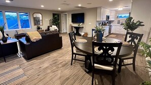 Una sala moderna con un sofá marrón oscuro, una mesa de comedor con sillas y una cocina con armarios blancos y un ventanal grande.