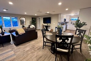 Una sala moderna con un sofá marrón oscuro, una mesa de comedor con sillas y una cocina con armarios blancos y un ventanal grande.