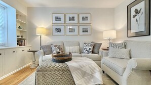 A living room with a sofa, ottoman, and a coffee table. There are framed pictures on the wall and a lamp on a side table. - Discover Tranquility at Carolina Cottage - Cozy 2-Bedroom Retreat in Hickory, NC (Hickory)