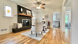A spacious living room with hardwood floors, a fireplace, a ceiling fan, and a view into the kitchen. - Discover Tranquility at Carolina Cottage - Cozy 2-Bedroom Retreat in Hickory, NC (Hickory)