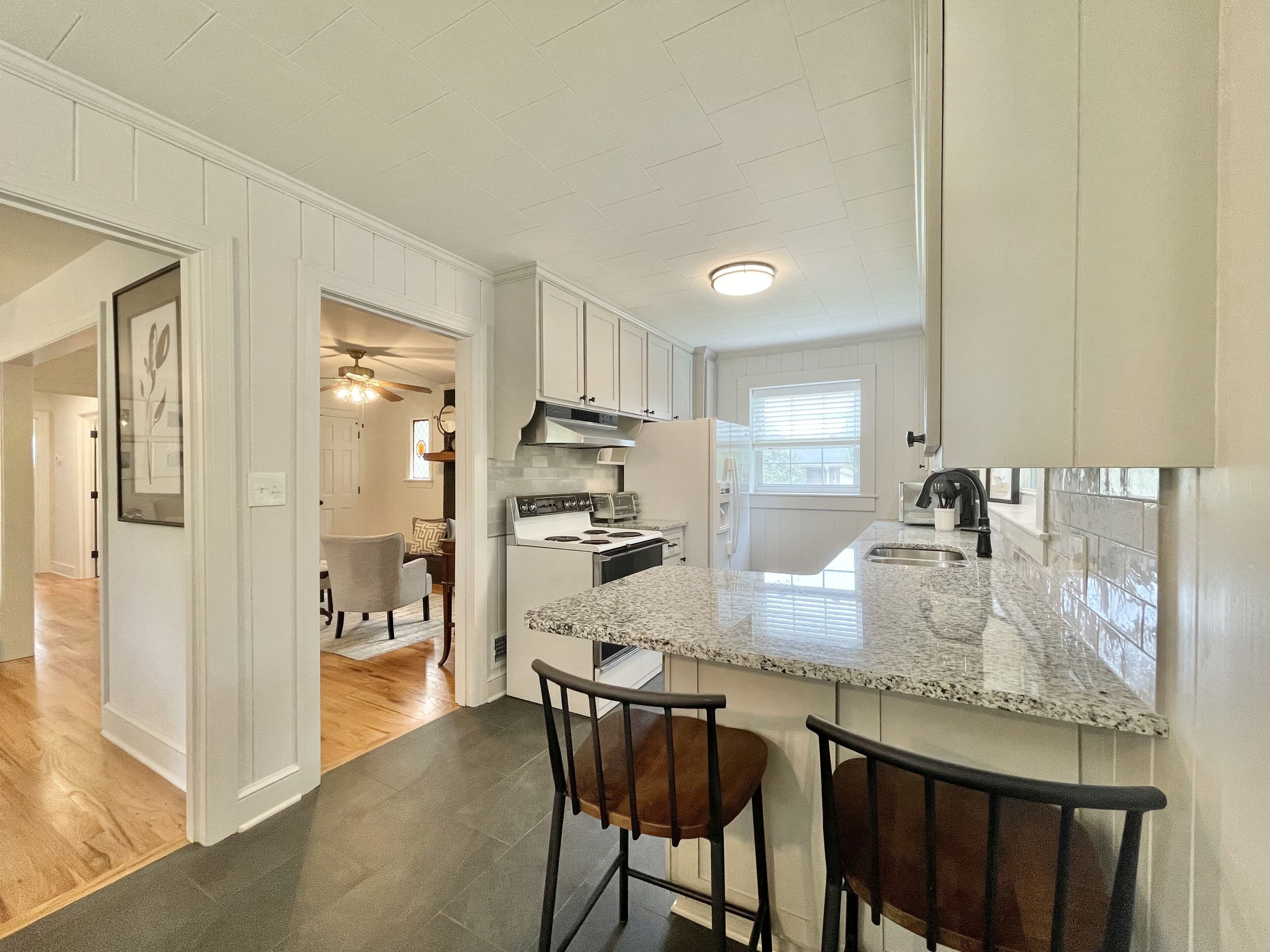 A kitchen with white cabinetry, a granite countertop, and a stainless steel range hood. There are two bar stools and a window providing natural light.