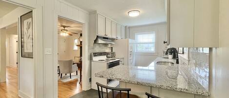 A kitchen with white cabinetry, a granite countertop, and a stainless steel range hood. There are two bar stools and a window providing natural light.