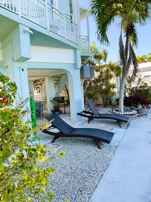 A modern outdoor lounge area with two black lounge chairs, a covered seating area, and a palm tree.