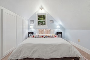 A bedroom with a sloped ceiling, a bed with a colorful quilt, two bedside lamps, and a window with a view of greenery. - Cozy Sapphire Retreat | Fireplace • Dog Friendly (Sapphire)