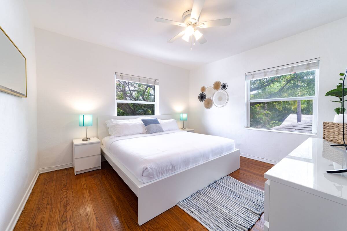 Une chambre moderne avec un grand lit, deux tables de chevet, un ventilateur de plafond et deux fenêtres équipées de stores.