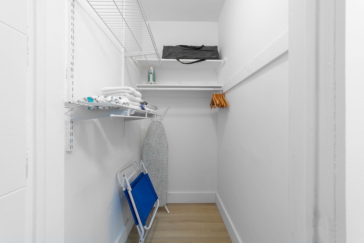 Un lavadero pequeño con paredes blancas, una estantería de metal con ropa doblada, un carro de lavandería azul y una plancha.
