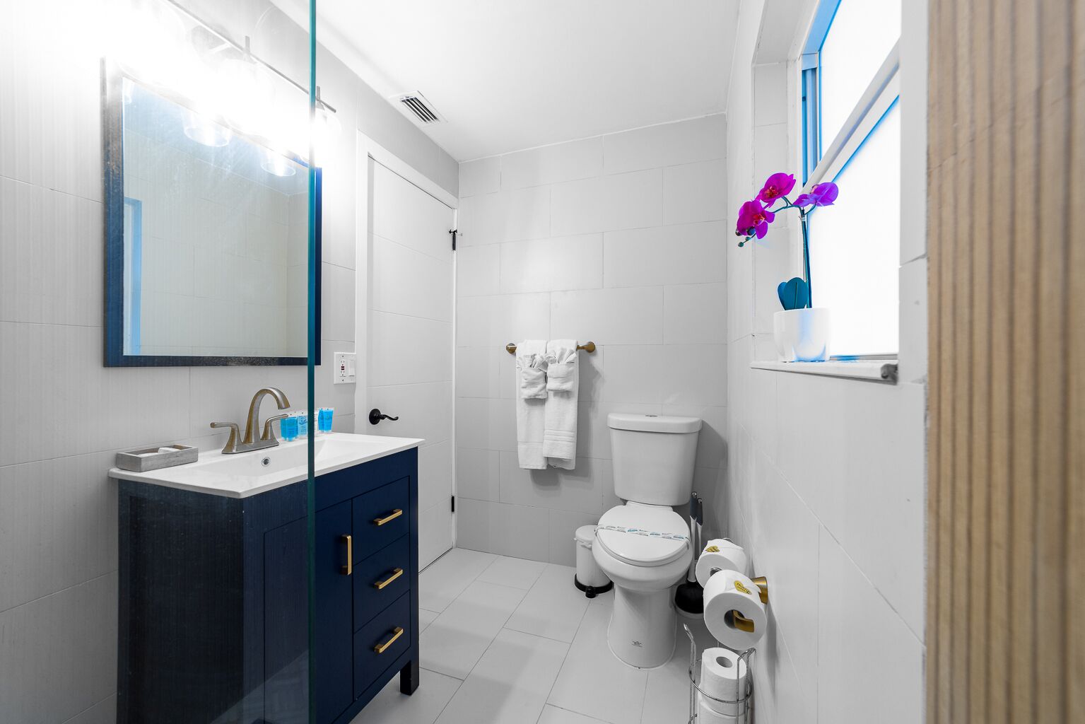 Un bagno moderno con un lavandino bianco, un mobile bagno blu scuro, un water bianco e una finestra con fiori viola.
