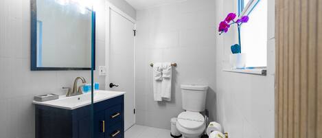 Un bagno moderno con un lavandino bianco, un mobile bagno blu scuro, un water bianco e una finestra con fiori viola.