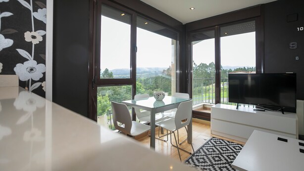 A modern living room with a glass dining table, white chairs, a flat-screen TV, and large windows offering a view of the outdoors. - Miralvalle Apartment (Valoria Cantabria)