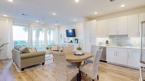 Un salon moderne avec un canapé, une table à manger et des chaises en osier. On y trouve également un coin cuisine avec des meubles blancs et un téléviseur fixé au mur.
