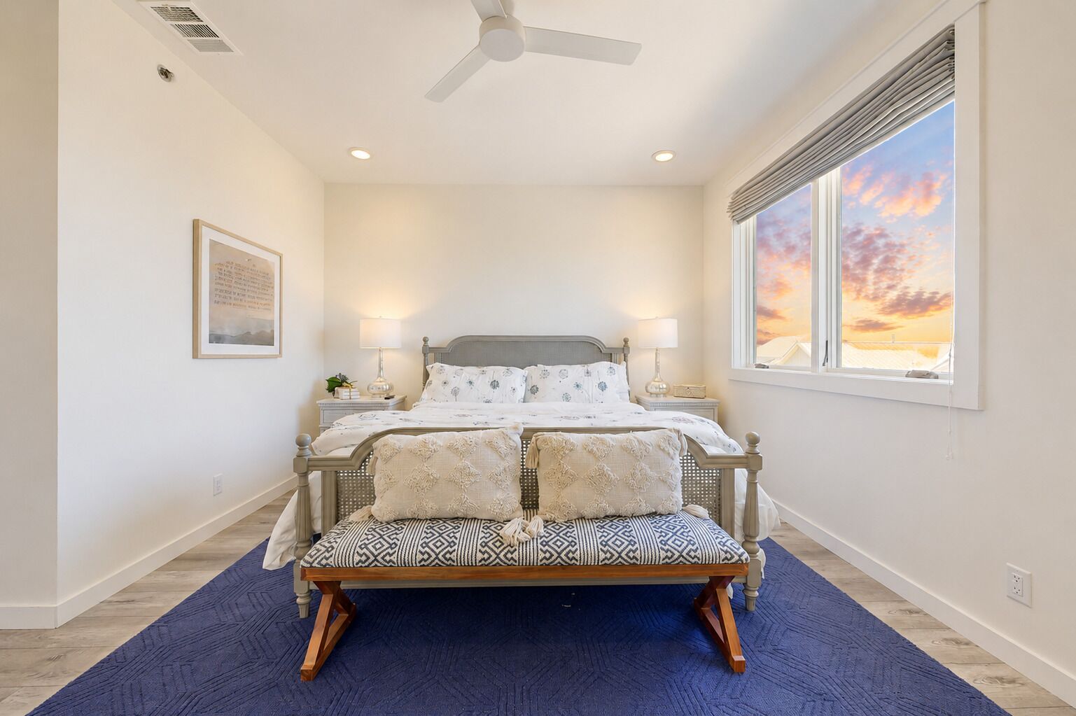 A bedroom with a bed, a bench, a ceiling fan, a window with a view, and a framed picture on the wall.