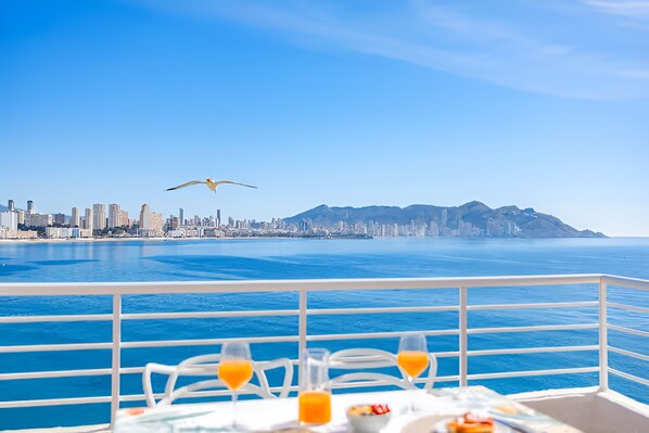 A table with two glasses of orange juice, a bowl of fruit, and a plate of food on a balcony overlooking a city skyline and the ocean.
