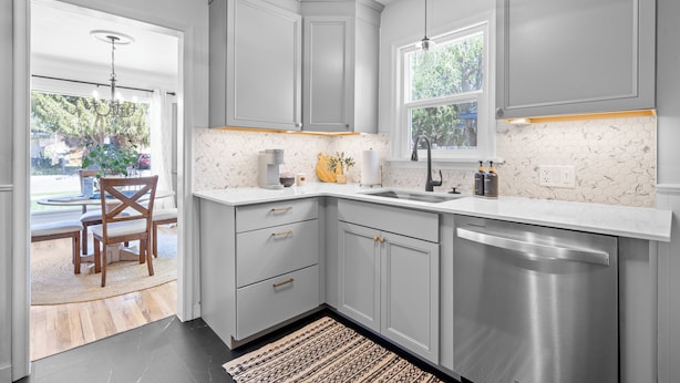 A kitchen with grey cabinets, a stainless steel dishwasher, a window with a view, and a wooden dining table with chairs. - Remodeled home with vintage charm. Centrally located near downtown Idaho Falls. (Idaho Falls)