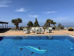 A swimming pool with lounge chairs and a sofa, surrounded by cacti and trees.