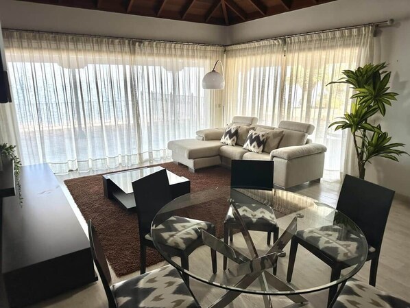 A modern living room with a glass table, two sofas, and a large window with sheer curtains.