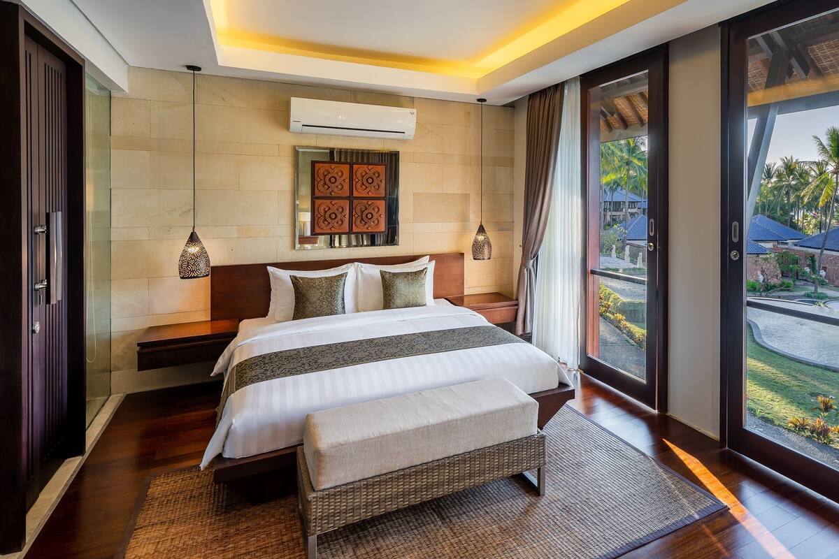 A modern bedroom with a large bed, a wall-mounted air conditioner, and a view of a landscaped area with palm trees.