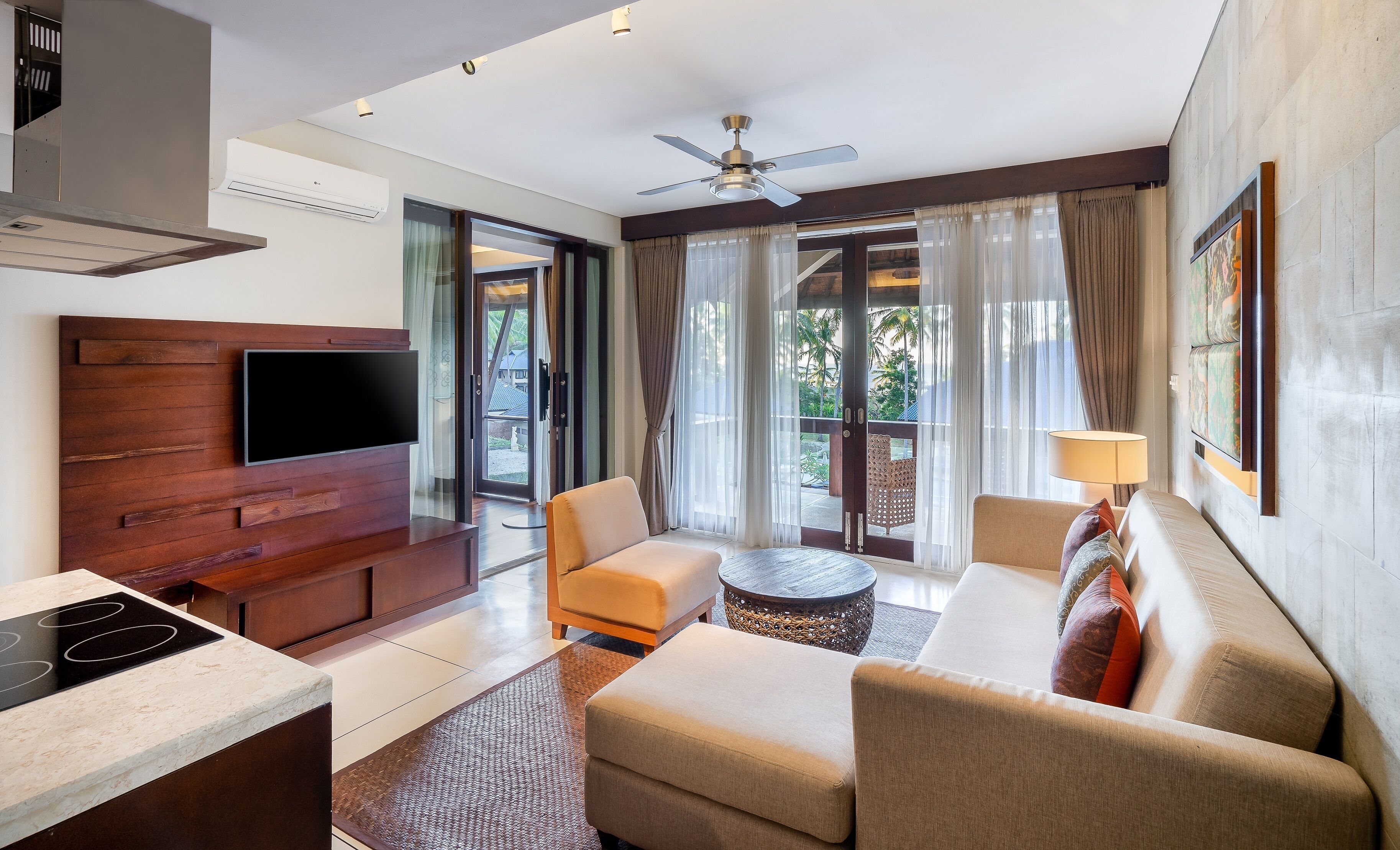 Una habitación de hotel moderna con televisor de pantalla plana, un panel de madera decorativo, ventilador de techo, sofá, mesa de centro y vistas al exterior.