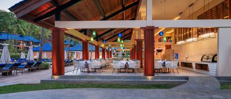 An outdoor dining area with tables and chairs, surrounded by palm trees and a pool.