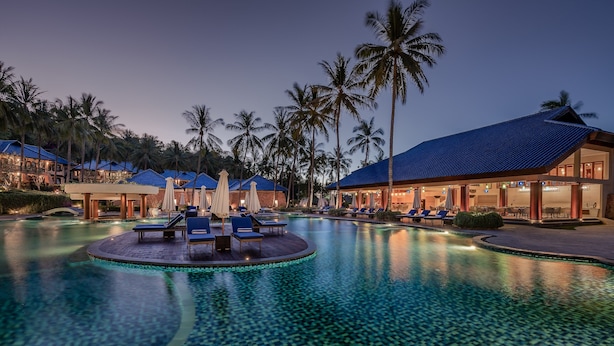 A resort pool area with lounge chairs and umbrellas, surrounded by palm trees and buildings with blue roofs. - Sundancer Residences & Villas Lombok (Sekotong Barat)