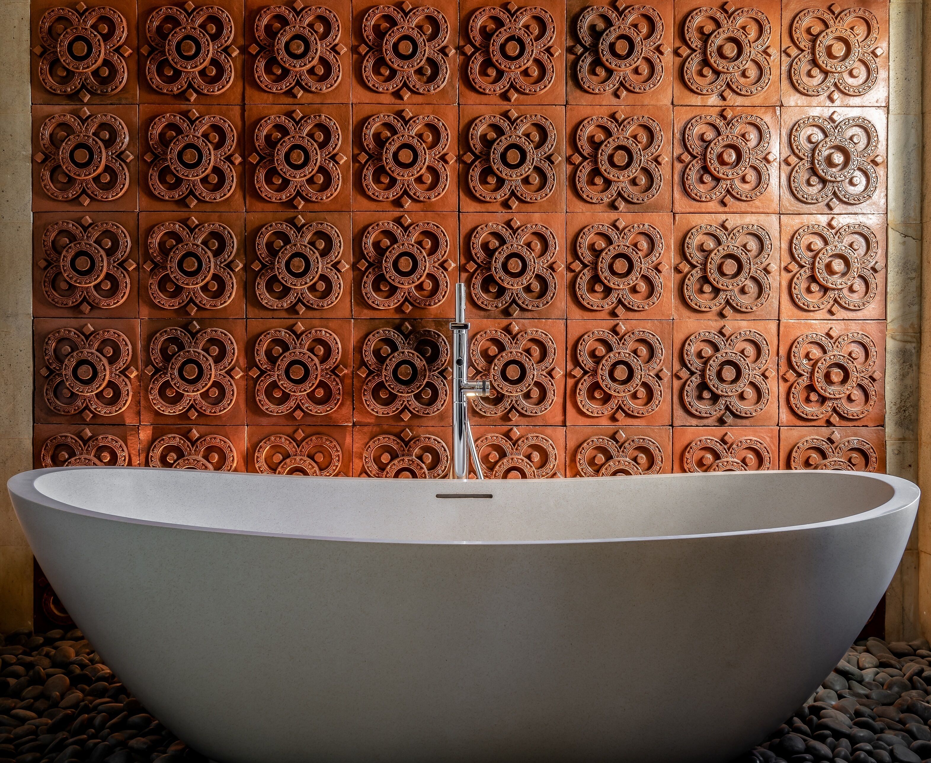 A modern bathroom with a freestanding bathtub, a tiled wall with intricate patterns, and a stone floor.
