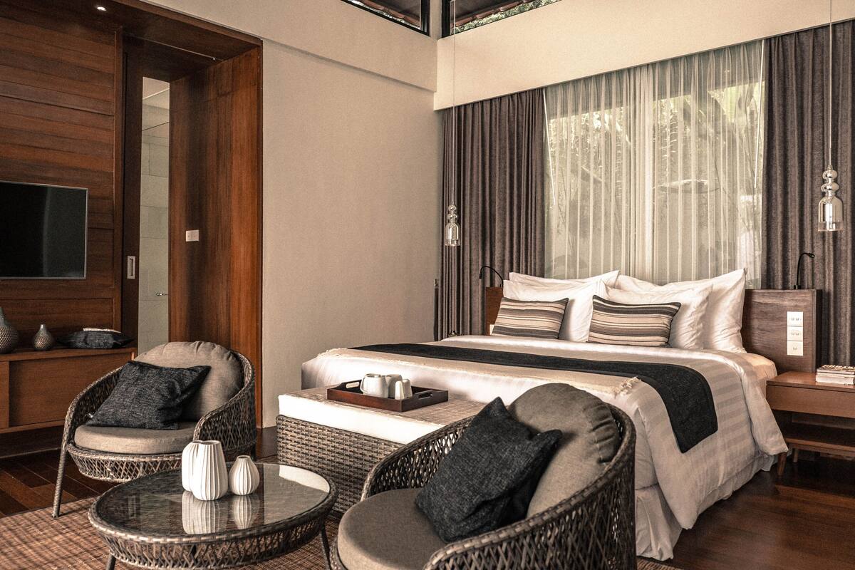 A modern hotel room with a large bed, two wicker chairs, a small table, and a TV on the left wall.