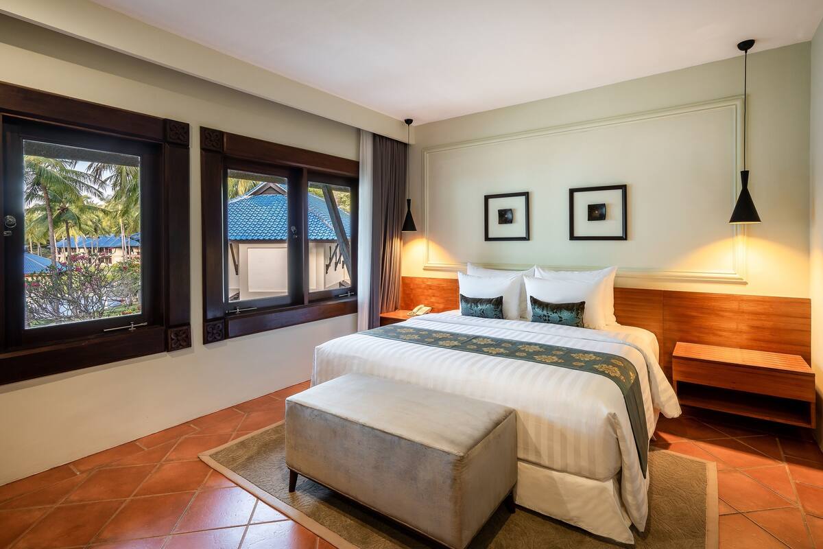 A hotel room with a large bed, a wooden nightstand, a bench, and a window with a view of palm trees.