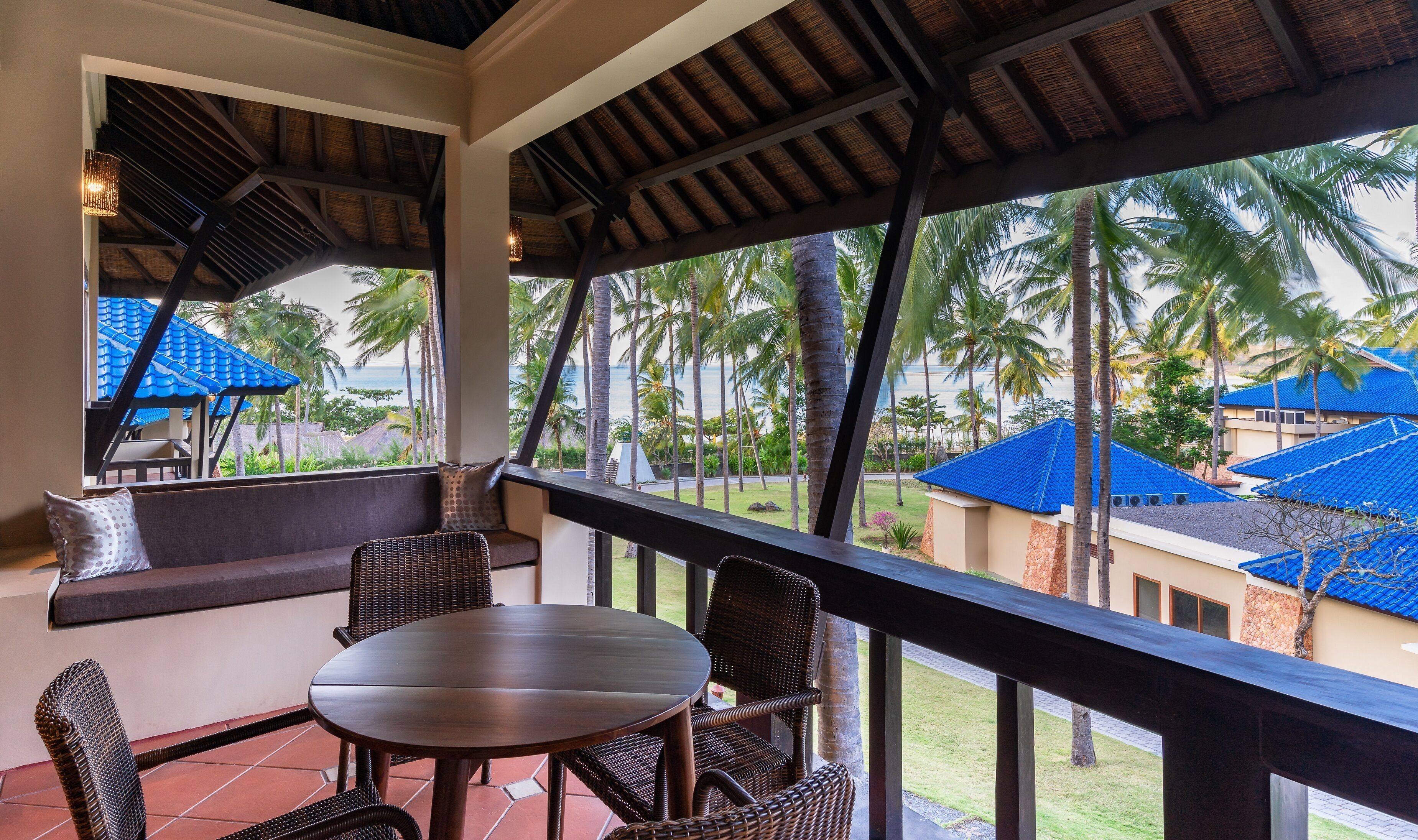 A covered outdoor seating area with wicker furniture and a table, overlooking a pool and palm trees.